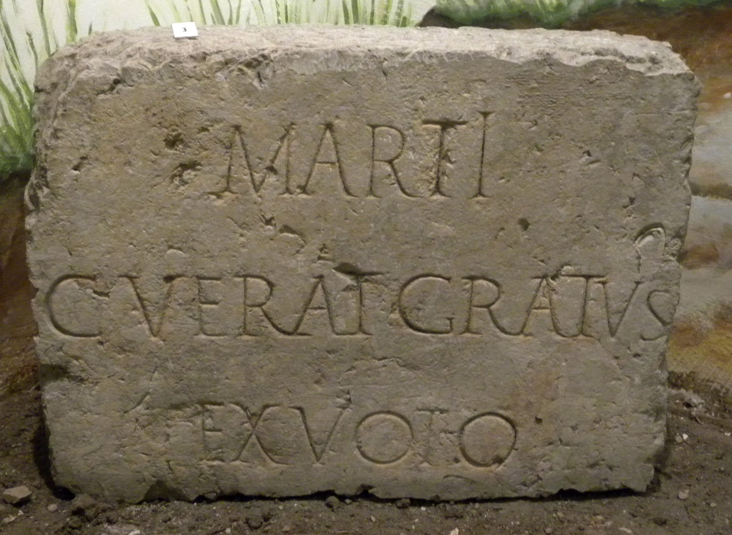 Bloc en pierre claire, rectangulaire. Un texte en latin est gravé en majuscule sur 3 lignes : MARTI / C. VERAT. GRATVS / EX VOTO