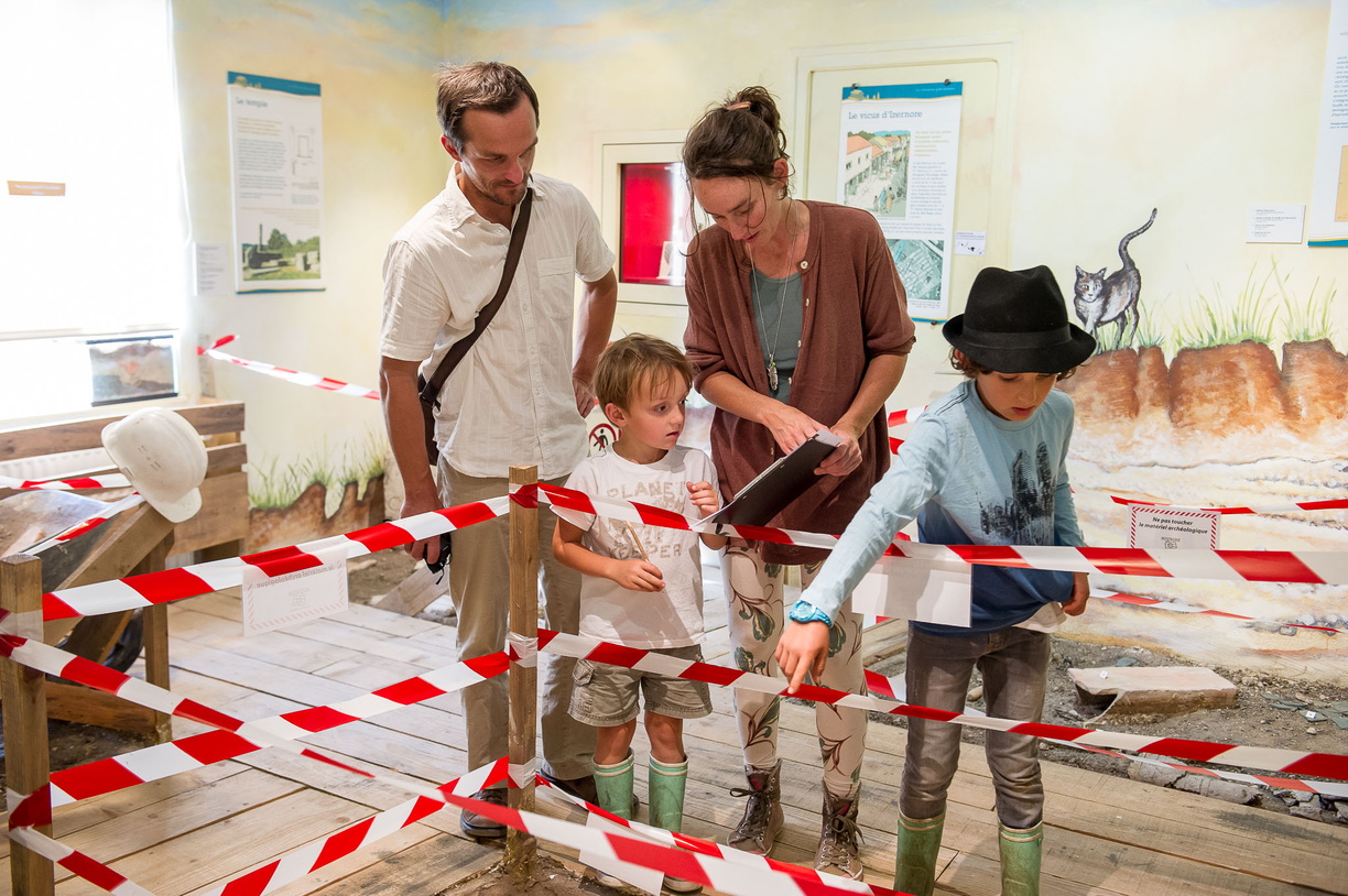 Visite du musée en famille. Les parents aident leurs enfants à compléter leur livret-jeu. Les enfants pointent du doigt les objets dans le musée.