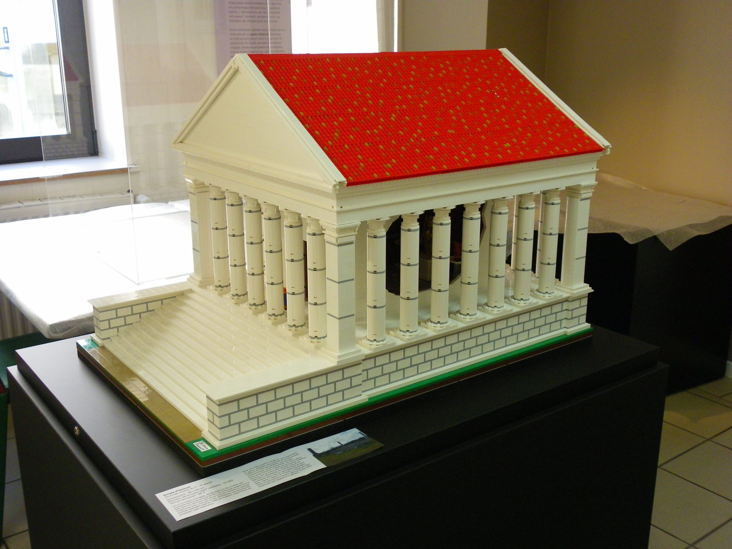 Maquettes en briques lego représentant le temple d'Izernore. Il est montré tel qu'il était il y a 2000 ans. L'escalier, les colonnes et piliers sont blancs. Le toit est rouge. On le voit de 3/4, orienté vers la gauche.